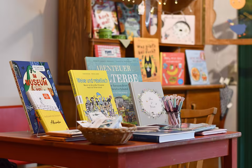 Bücher stehen auf einem roten Tisch, im Hintergrund ist unscharf eine Holzkommode mit Büchern darauf